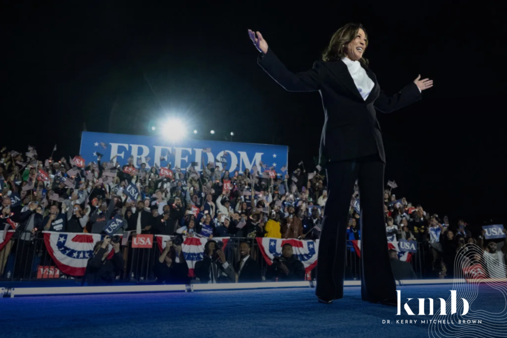 Kamala Harris standing on stage at a political rally, smiling and gesturing to a crowd holding American flags and banners with the word 'FREEDOM' displayed behind her. The logo for Dr. Kerry Mitchell Brown is visible in the bottom right corner